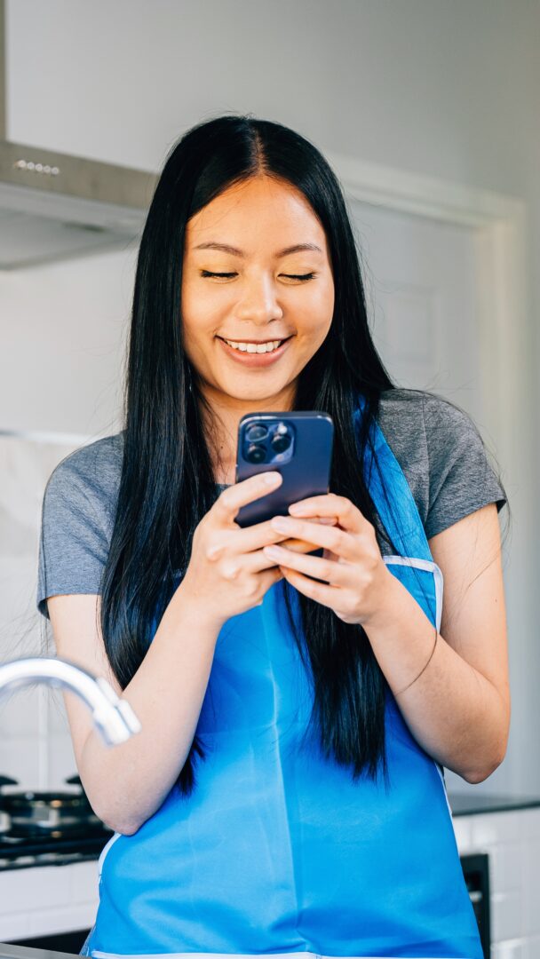 Smiling woman reads messages on her cellphone leaning on the kitchen